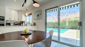 Cocina moderna con gabinetes blancos, una mesa de comedor con sillas y vista a una piscina y montañas a través de grandes puertas correderas de vidrio.
