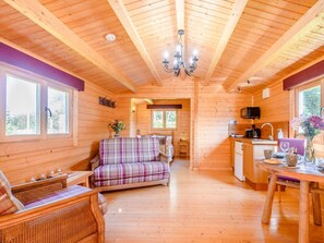 A cabin interior with a wooden floor, a plaid sofa, a dining table, a kitchen area, and a chandelier. - 1 Bed in Helensburgh (oc-78989) (Portincaple)
