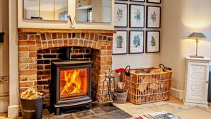 A fireplace with a brick surround, a wooden crate with logs, and framed botanical prints on the wall. - 3 Bed in Langport (oc-94302) (Curry Rivel)