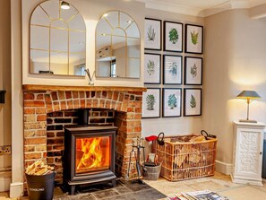 A fireplace with a brick surround, a wooden crate with logs, and framed botanical prints on the wall. - 3 Bed in Langport (oc-94302) (Curry Rivel)