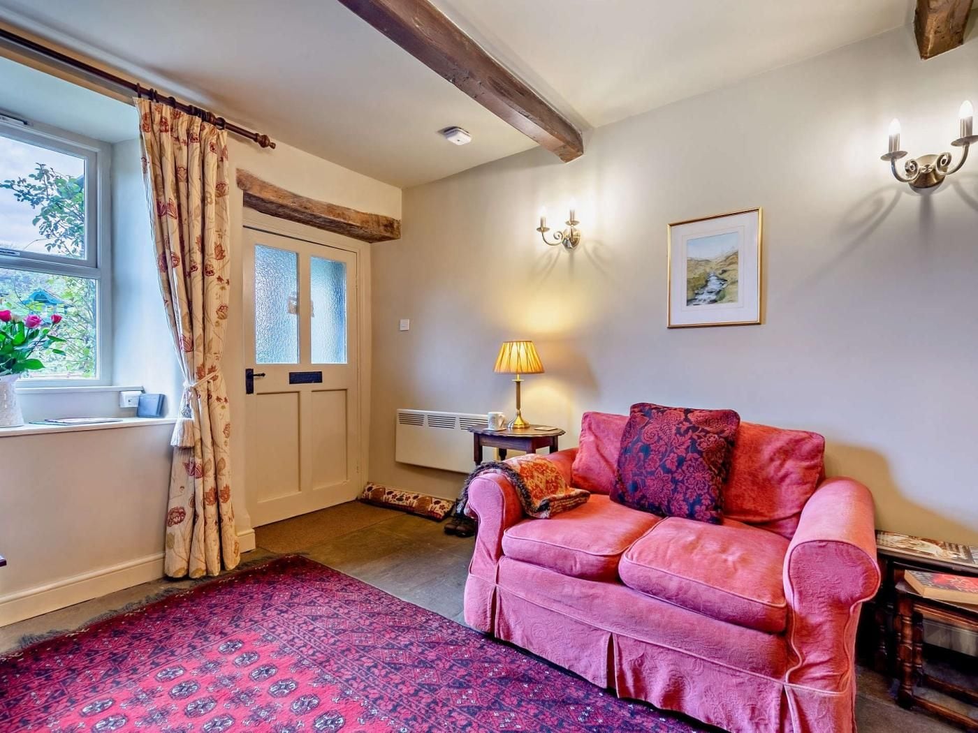 A cozy room with a pink sofa, a patterned rug, a window with curtains, and a wall-mounted lamp.