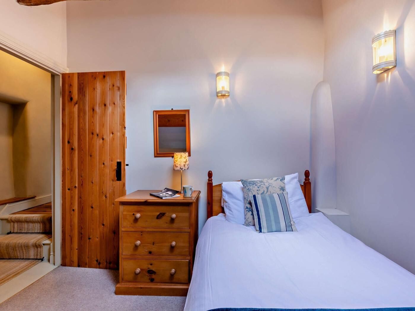 Une chambre avec une commode en bois, un lit avec une literie blanche, une porte en bois et deux appliques murales.