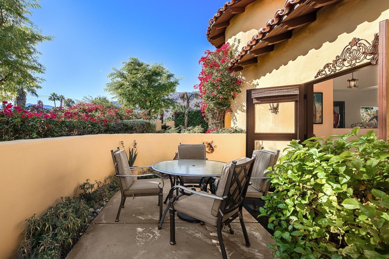 An outdoor patio with a round table and chairs, surrounded by plants and a building with a tiled roof.