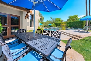 An outdoor patio with a table and chairs, a pool, and a blue umbrella. - Outdoor Shower, Swim-Up Wetbar, Hot Tub, Putting Area, Firepit, EV Charger (Palm Springs)