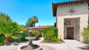 A courtyard with a stone fountain, palm trees, and a building with a tiled roof and a star-shaped decoration. - Outdoor Shower, Swim-Up Wetbar, Hot Tub, Putting Area, Firepit, EV Charger (Palm Springs)