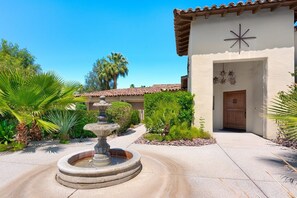A courtyard with a stone fountain, palm trees, and a building with a tiled roof and a star-shaped decoration. - Outdoor Shower, Swim-Up Wetbar, Hot Tub, Putting Area, Firepit, EV Charger (Palm Springs)