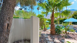A stone water feature with a cascading waterfall, surrounded by pebbles and palm trees, with a pool and lounge area in the background. - Outdoor Shower, Swim-Up Wetbar, Hot Tub, Putting Area, Firepit, EV Charger (Palm Springs)