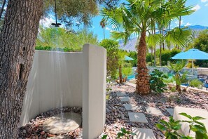 A stone water feature with a cascading waterfall, surrounded by pebbles and palm trees, with a pool and lounge area in the background. - Outdoor Shower, Swim-Up Wetbar, Hot Tub, Putting Area, Firepit, EV Charger (Palm Springs)