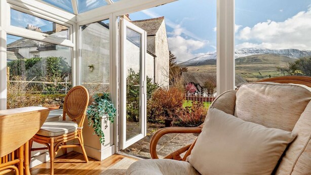 A conservatory with a view of a rural landscape, a dining table, chairs, and a sofa. - 3 Bed in Keswick (oc-94346) (Threlkeld)