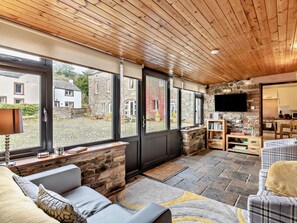 A cozy interior with a stone fireplace, a flat-screen TV mounted on a stone wall, and a view of the outside through large windows. - 2 Bed in Bassenthwaite (oc-83287) (Bassenthwaite)