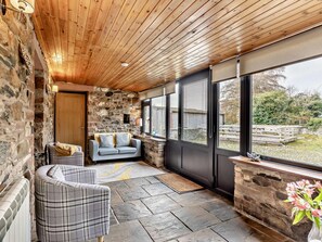 A cozy room with a stone wall, a wooden ceiling, a grey sofa, a plaid armchair, and a large window with a view of a garden. - 2 Bed in Bassenthwaite (oc-83287) (Bassenthwaite)