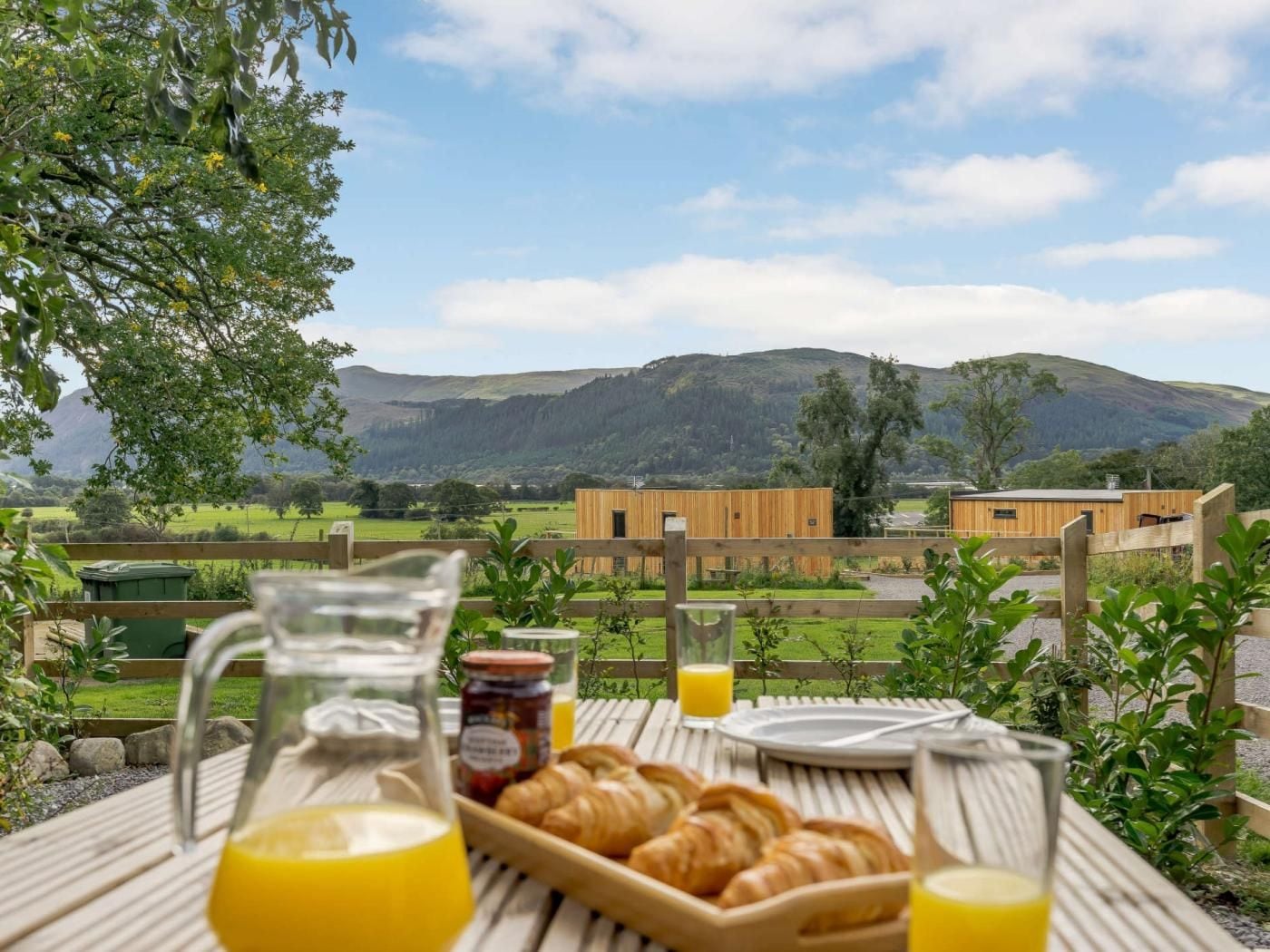 A breakfast setup with croissants and orange juice on a wooden table, overlooking a scenic landscape with hills and a building.
