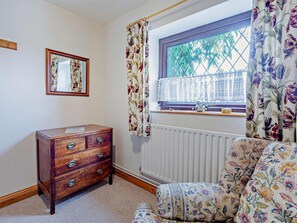 Une chambre avec une commode en bois, une fenêtre ornée de rideaux à motifs floraux, un radiateur et un miroir accroché au mur.