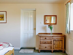 Une pièce avec une porte blanche, une commode en bois dotée de tiroirs, un miroir au-dessus, et un lit recouvert d’une couvre-lit à motifs.