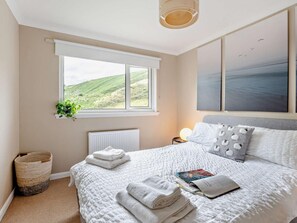 A bedroom with a large window, a radiator, a bedside table with a lamp, a bed with white bedding, and a wall with two framed pictures. - 5 Bed in Dunbar (oc-94334) (Dunbar)