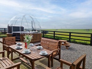 Een buitenterras met houten tafels en banken, een overdekte barbecue, en een uitzicht op windturbines in de verte.