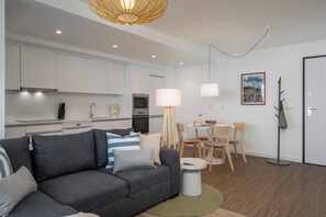 Un salon moderne avec un canapé gris, une table à manger avec des chaises, un coin cuisine doté de meubles blancs et un luminaire suspendu.
