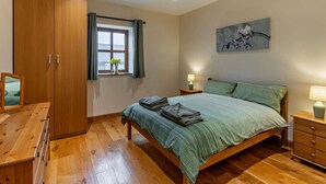 Ein Schlafzimmer mit Holzschrank, einem Bett mit grüner Bettdecke, einem Nachttisch mit Lampe und einem Fenster mit Vorhängen.