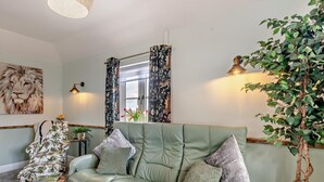 Un sofá verde con almohadones, un sillón floral, un cuadro de un león, una planta en maceta y una ventana con cortinas.