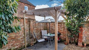 Un patio con una pérgola de madera, una mesa redonda y sillas, rodeado de un muro de ladrillo y follaje.
