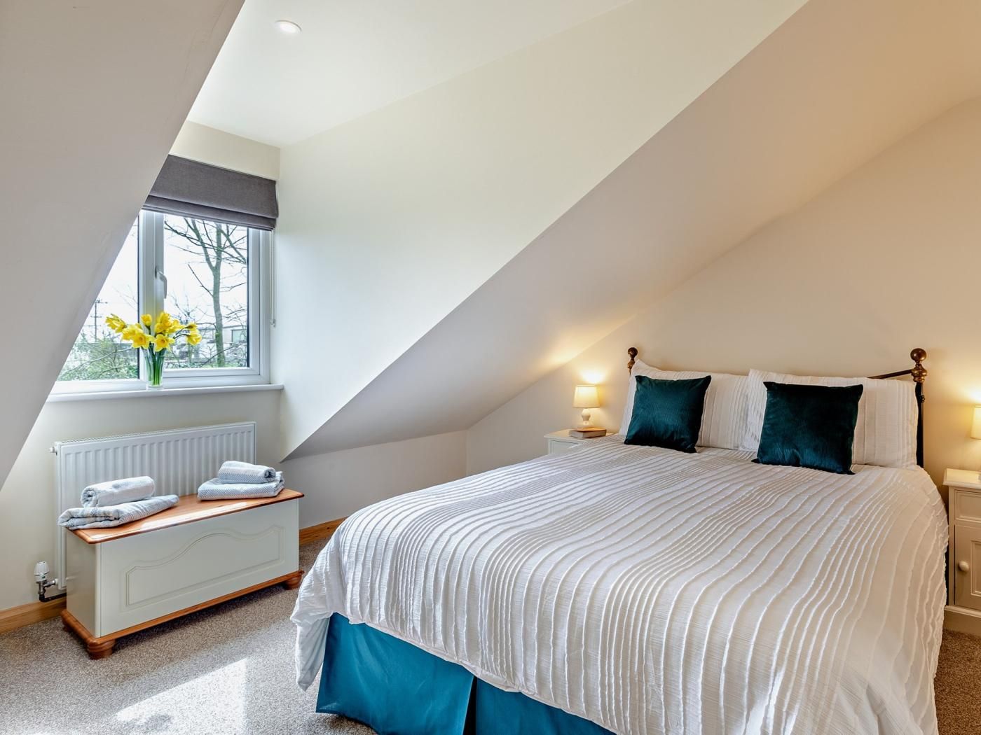 Une chambre avec un plafond en pente, un grand lit avec une literie blanche, deux lampes de chevet, une fenêtre donnant sur des arbres et un radiateur.
