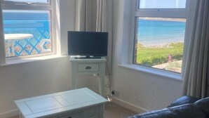 Una habitación con televisión sobre un mueble bajo, una mesita, un sofá y una ventana con vista a la playa.