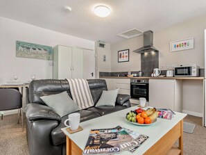 A modern living room with a leather sofa, a coffee table with magazines, and a kitchen area with a microwave and oven. - 1 Bed in Woolacombe (oc-76348) (Woolacombe)