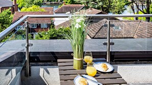 En tagterrasse med et bord dækket til morgenmad, inklusive croissanter og juice, med udsigt over et boligområde med træer og huse.