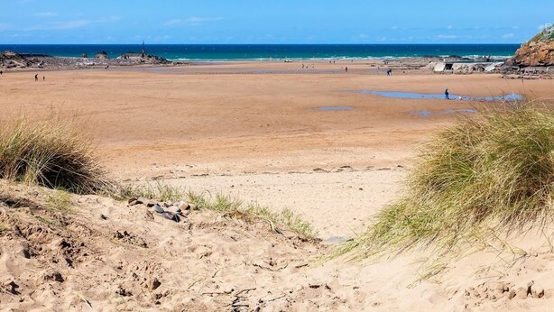 Ein weiter Sandstrand mit blauem Himmel, Felsformationen und Blick auf das Meer in der Ferne.