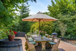 An outdoor dining area with a table set for six, surrounded by wooden chairs, under a large umbrella.