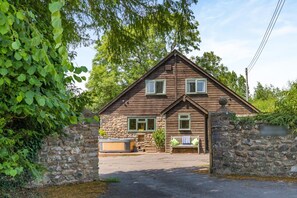 A two-story wooden house with a stone wall entrance, a bench outside, and a hot tub in the driveway.