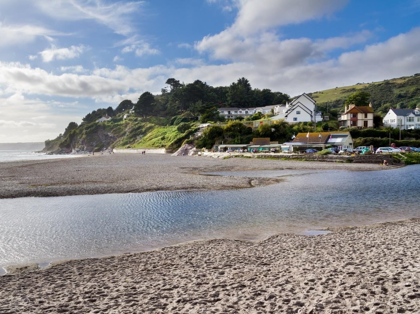 A coastal beach with houses on a hillside, a sandy foreground, and a calm sea.