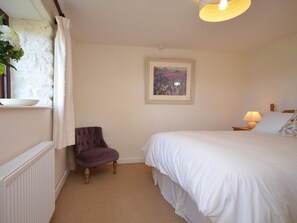 Quarto com cama, poltrona, radiador, janela com cortinas, quadro na parede e abajur na mesa de cabeceira.