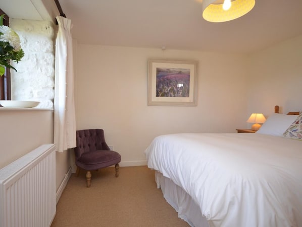 A bedroom with a bed, a chair, a radiator, a window with curtains, a painting on the wall, and a lamp on the bedside table.