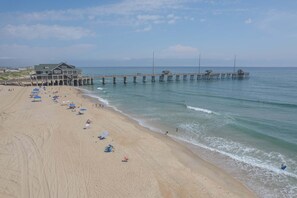 A beach with a pier extending into the water, a large building on the left, and people on the sand. - 5726 - Ellington Cottage By The Sea, Oceanfront retreat with hot tub, decks, and ocean views. (Nags Head)