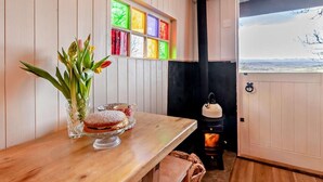 Ein Holzinnenraum mit einem Tisch, auf dem sich eine Blumenvase und ein Dessert befinden, ein farbenfrohes Fenster und ein Kaminofen.