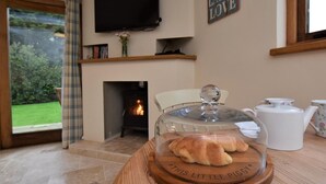 A cozy living room with a fireplace, a television, and a table with a glass dome displaying croissants. - 1 Bed in Axminster (oc-36491) (Dalwood)