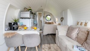 A compact, well-lit room with a kitchenette, a dining area with a checkered table, and a cozy seating area with a sofa and cushions.