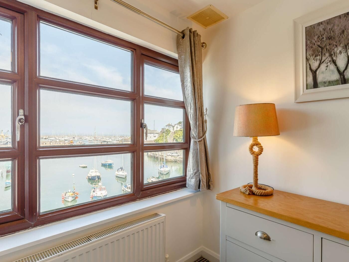A room with a bay window overlooking a marina, a radiator below the window, a wooden dresser with a lamp, and a framed picture on the wall.