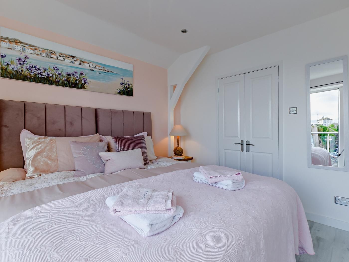 A bedroom with a large bed, a headboard, a painting above the bed, a bedside table with a lamp, and a window with a view.