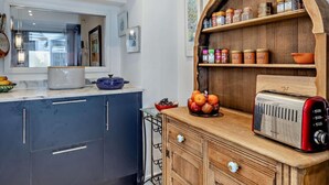 Una cocina con estante de madera que contiene frascos, un tostador y un cuenco de fruta.