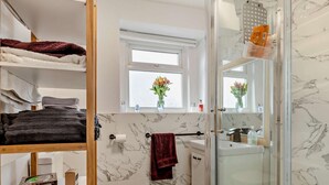Baño con inodoro, lavamanos y ducha. Hay toallas y artículos de aseo en estantes.