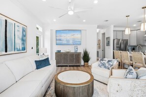 A modern living room with a white sofa, a round coffee table, and a dining area with wicker chairs. - Serenity Seas - All New Luxury Home Across from Beach w/ Heated Pool, Mini Golf (Indian Rocks Beach)