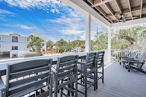 A covered outdoor seating area with wooden chairs and tables, overlooking a residential street with houses and palm trees. - Serenity Seas - All New Luxury Home Across from Beach w/ Heated Pool, Mini Golf (Indian Rocks Beach)