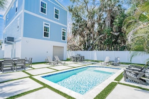 A pool with a geometric tile pattern, surrounded by a lawn with a white fence, and a building with blue siding and multiple windows. - Serenity Seas - All New Luxury Home Across from Beach w/ Heated Pool, Mini Golf (Indian Rocks Beach)