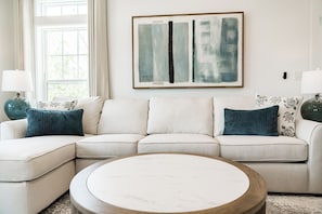A living room with a sectional sofa, a round coffee table, and a large abstract painting on the wall. - Serenity Seas - All New Luxury Home Across from Beach w/ Heated Pool, Mini Golf (Indian Rocks Beach)