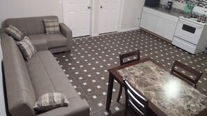 Ein Wohnzimmer mit einer Couch, einem Esstisch mit Stühlen, einer Küchenzeile mit Schränken und Geräten sowie einem gemusterten Boden.