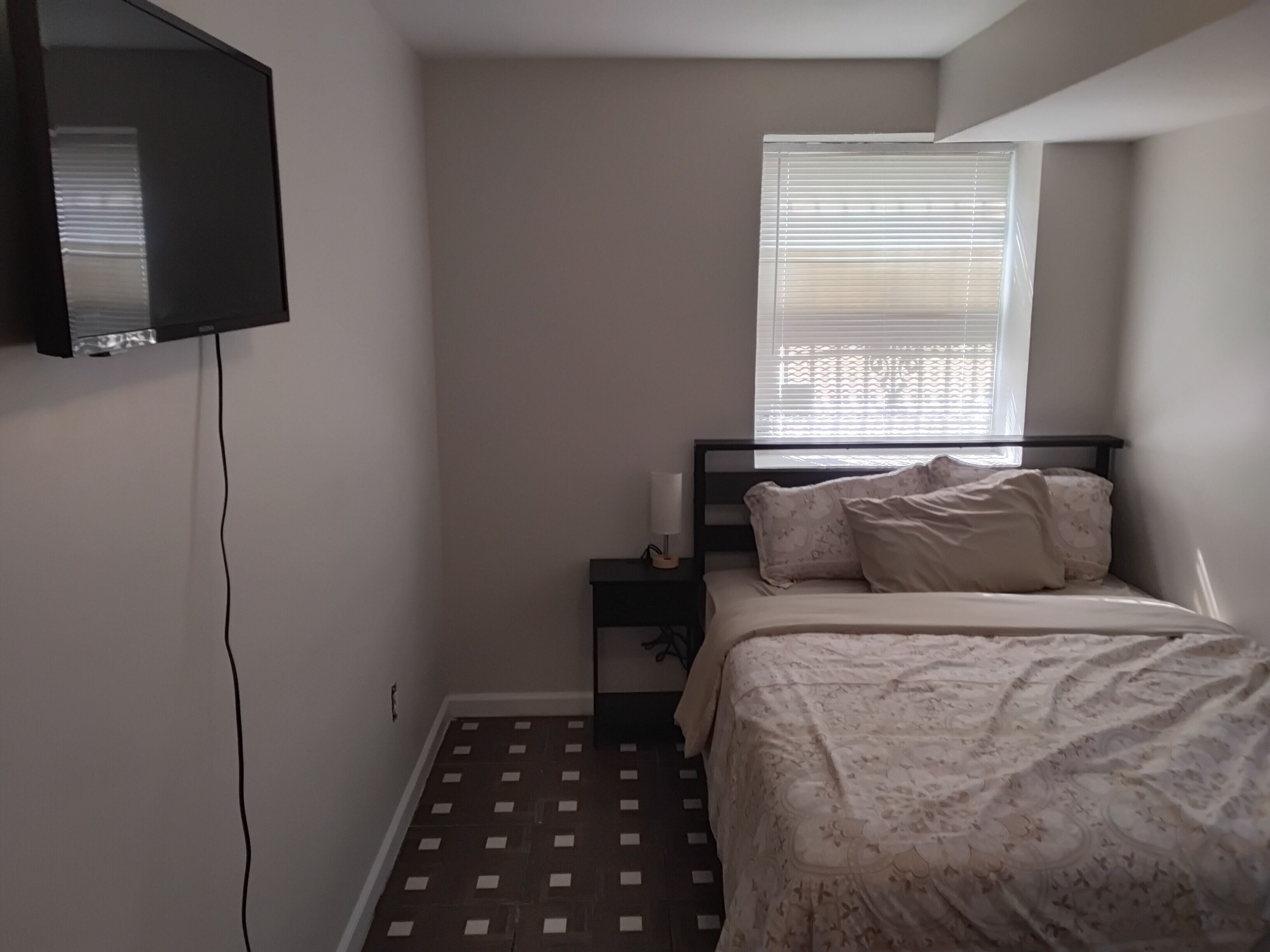 A bedroom with a bed, a nightstand, a TV mounted on the wall, and a window with blinds.