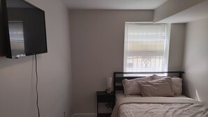 Ein Schlafzimmer mit einem Bett, einem Nachttisch, einem an der Wand befestigten Fernseher und einem Fenster mit Jalousien.