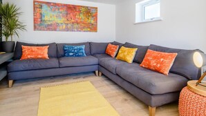 Ein modernes Wohnzimmer mit einem grauen Ecksofa, bunten Kissen, einem großen abstrakten Gemälde, einer Topfpflanze und einem gelben Teppich.